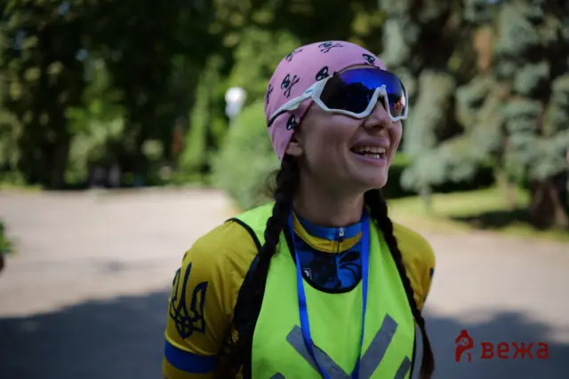 Smiling person wearing a neon safety vest, pink bandana, and white reflective sunglasses outdoors in a park.