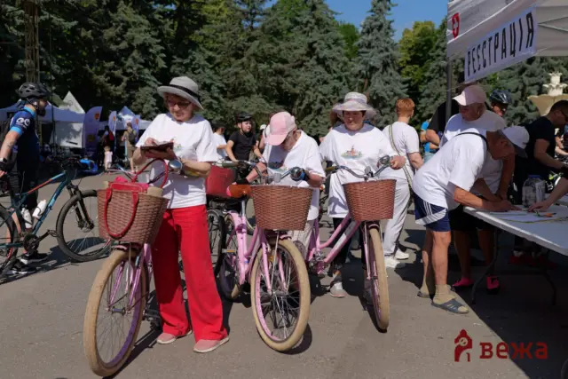 Group of people with bicycles and wicker baskets at an outdoor event, some wearing white T-shirts and hats near a registration tent.