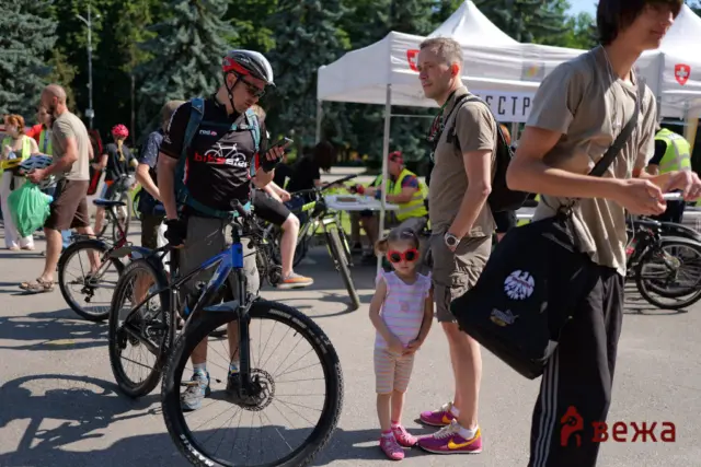 People with bicycles at an outdoor event, children and adults mingle near a white canopy tent with a red emblem in a sunny park setting.