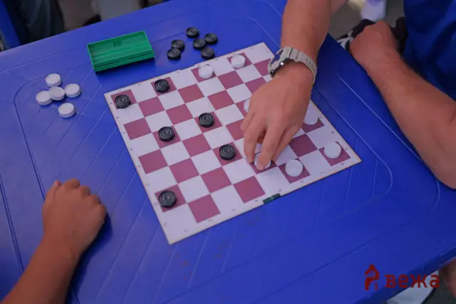 Person making a move in a checkers game on a pink-and-white board; black and white discs scattered with a green storage tray on a blue table nearby