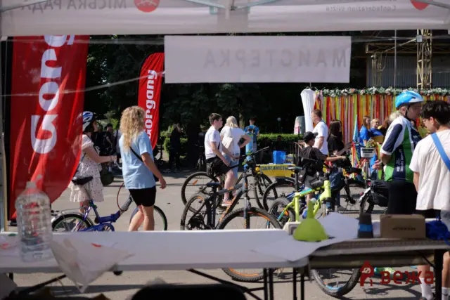 Outdoor bike event with many riders and colorful banners; people gather near a booth with a water bottle on a table in the foreground.