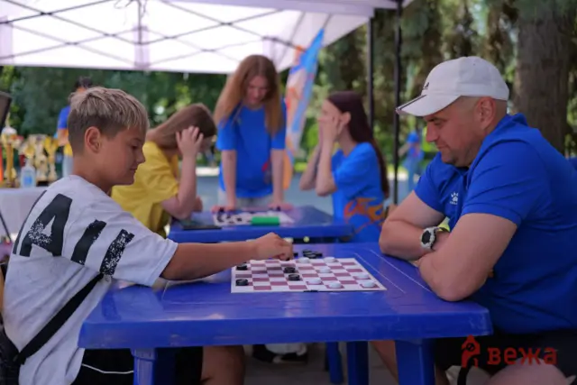 Boy in a white shirt moves a checker on a checkers board, facing a man in a white cap and blue shirt at an outdoor table under a canopy.