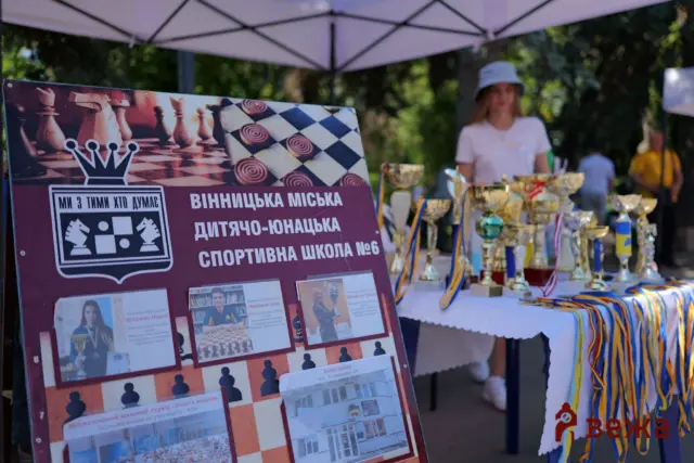 Maroon banner with a chessboard graphic and Ukrainian text about a city children’s sports school, with trophies on a table nearby under a canopy.