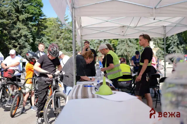 People with bicycles under a white canopy at a bike event; volunteers check-in and talk to riders in bright safety vests.