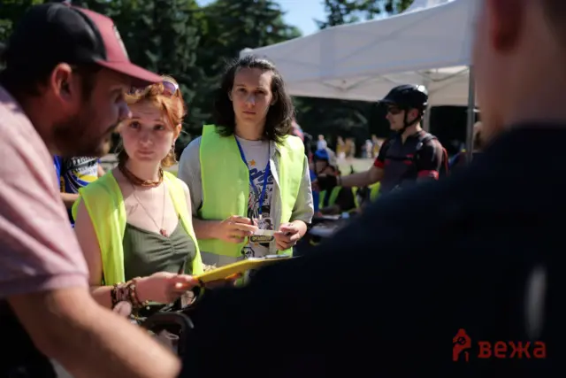 Young woman in a neon safety vest holding papers at an outdoor event, with volunteers and a tent in the background.