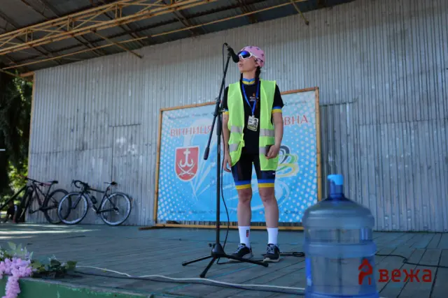 Person on an outdoor stage wearing a neon safety vest and pink bandana, speaking into a microphone.