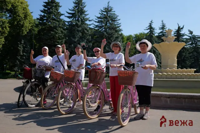 Group of people on pink bicycles waving in a sunny park near a large fountain and green trees.