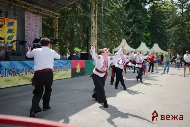 Dancers in traditional Ukrainian embroidered shirts perform on an outdoor stage as a crowd watches in a park festival.