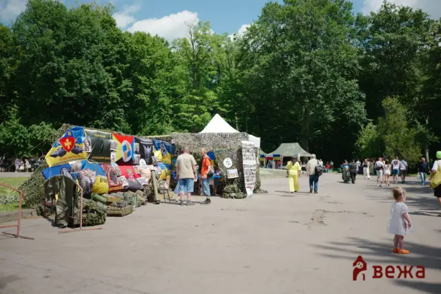 Outdoor market stalls with colorful banners and camouflage gear on display under a tree-lined park on a sunny day.