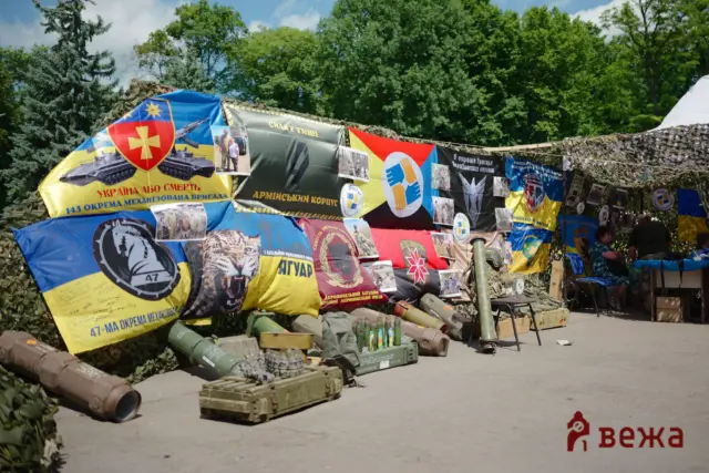Outdoor military exhibition with colorful banners, camouflage netting, and model tanks and missiles on the ground