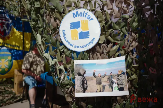 Round sign with three hands in blue and yellow joining together, surrounded by camouflage netting; edge text in Cyrillic suggests volunteering or aid messaging.