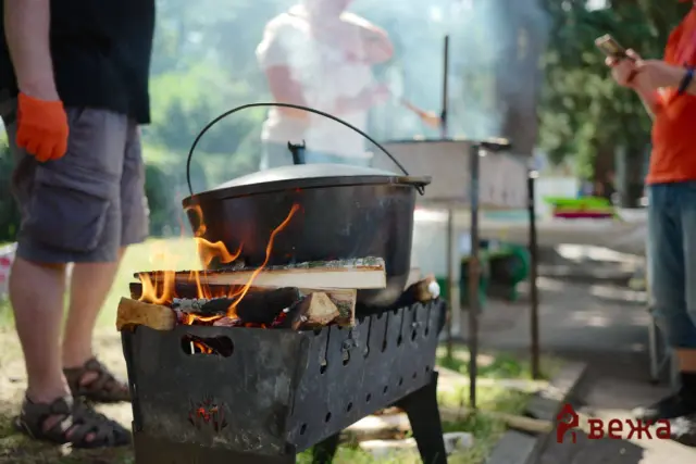 Outdoor barbecue scene with a large pot on a charcoal grill, flames licking the wood under the pot, and people standing nearby wearing casual clothes and orange gloves on one hand.