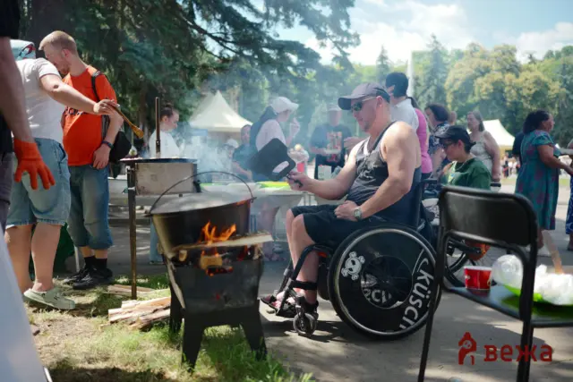 Man in a wheelchair grilling over an open fire while others mingle at an outdoor park barbecue.