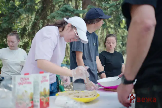 People standing around a table outdoors, wearing gloves and serving or arranging food while others watch, with trees in the background.