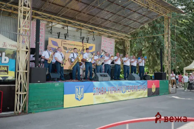 Brass band performing on an outdoor stage with blue-yellow banners and trees in the background.