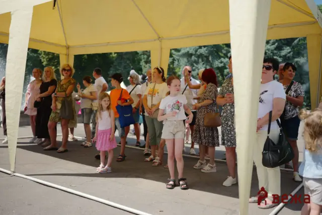 Group of people (men, women, and children) standing under a yellow event tent outdoors, waiting or watching something.
