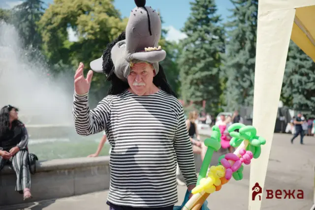 Man wearing a large plush wolf head hat and black-and-white striped shirt waves at the camera at an outdoor festival, with colorful balloon sculptures nearby.