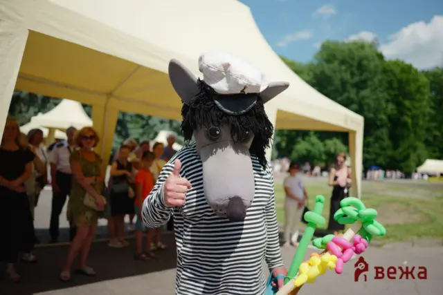 Person in a horse head mask and striped shirt giving a thumbs up at an outdoor festival with tents and balloons in view