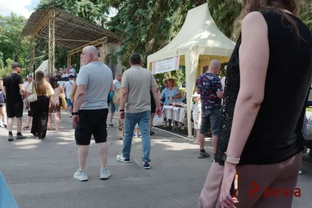 Crowd at an outdoor festival with a stage, tents, and trees on a sunny day, people walking and browsing stalls.