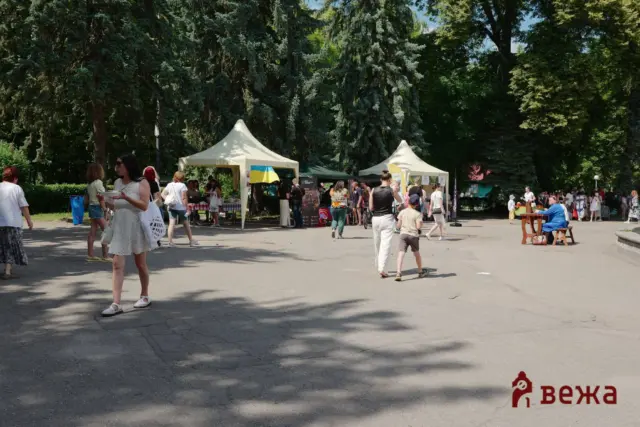 Outdoor market in a park with white tents, people browsing stalls, and tall trees overhead; a Ukrainian flag is visible near a stall and a few attendees stroll along the walkway.