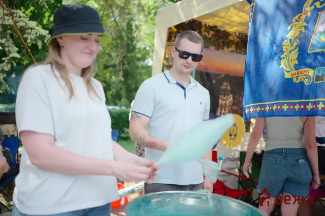 People at an outdoor booth blowing bubbles; a man wearing sunglasses and a light polo watches while a woman in a white shirt and black hat interacts nearby under a tent with a blue banner to the side.