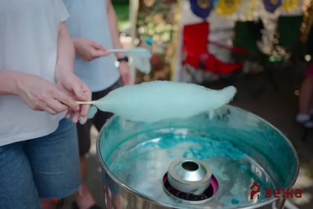 Hands spin turquoise cotton candy on sticks over a cotton candy machine at a fair or carnival setup, with blurred background stalls.