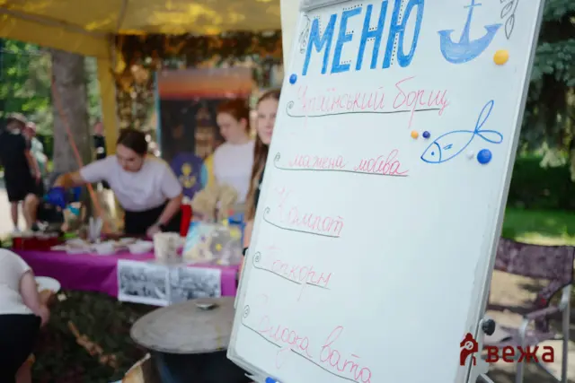 Outdoor market scene with a whiteboard sign in Ukrainian and a blue fish drawing; blurred shoppers in the background.