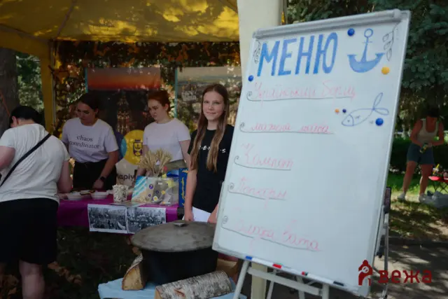 Outdoor festival booth with four people behind a table under a yellow canopy; a whiteboard menu stands to the right with handwritten Ukrainian items and drawings.