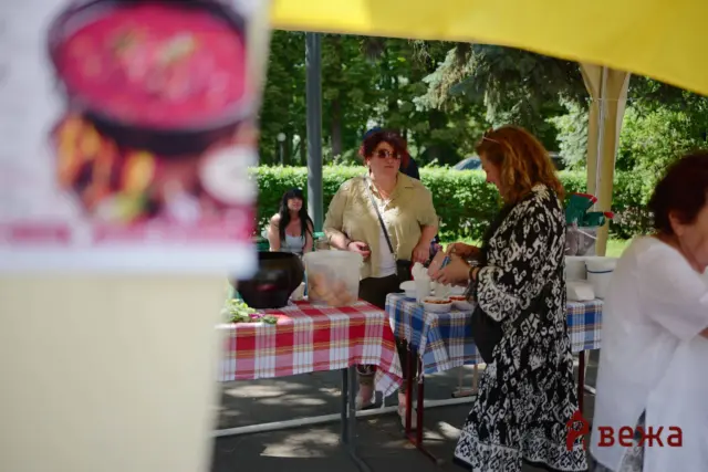 Outdoor market scene with people at a food stall under a yellow canopy; red and white and blue checkered tables hold bowls and containers, greenery in the background.