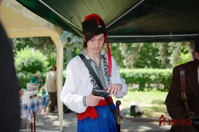 Young man in a traditional embroidered shirt with a red sash and black hat, holding a handheld recording device under a shade canopy at an outdoor event, focused on the device.