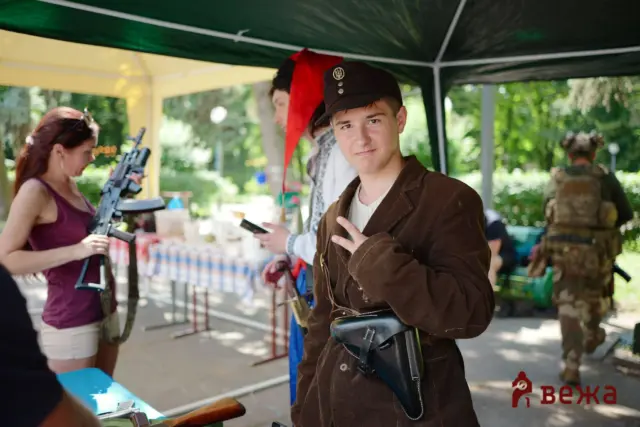 Young man in a vintage brown jacket and a dark cap, making a peace sign at an outdoor event under a green canopy.