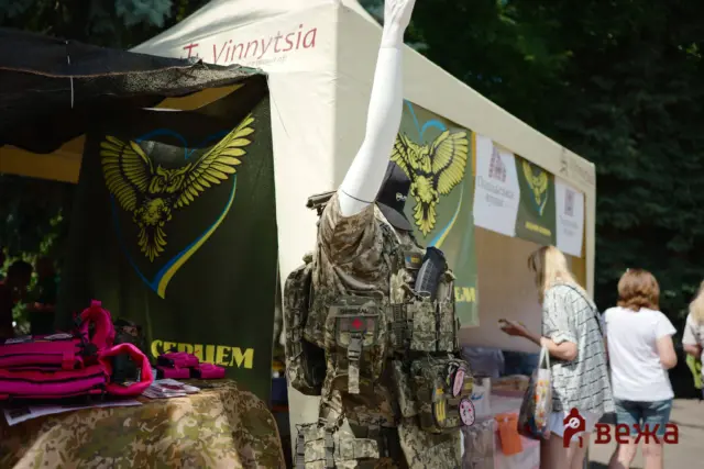Military display at an outdoor booth: mannequin in camo gear and tactical vest next to banners with a yellow eagle emblem and pink bags on a camo tablecloth.