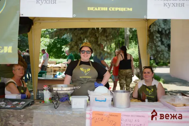 Smiling woman in apron at an outdoor food stall, with pots, trays, and other volunteers around under a canopy.