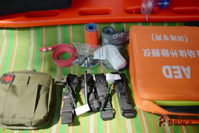 Orange medical rescue bag with white text sits on a striped mat, surrounded by rolled straps, cables, and tactical gear for emergency care/evacuation.