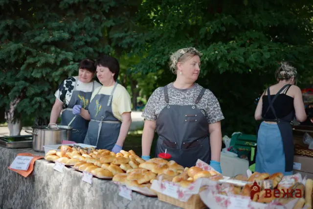 Three women in gray aprons sell fresh buns at an outdoor market, with a table of baked goods in front.