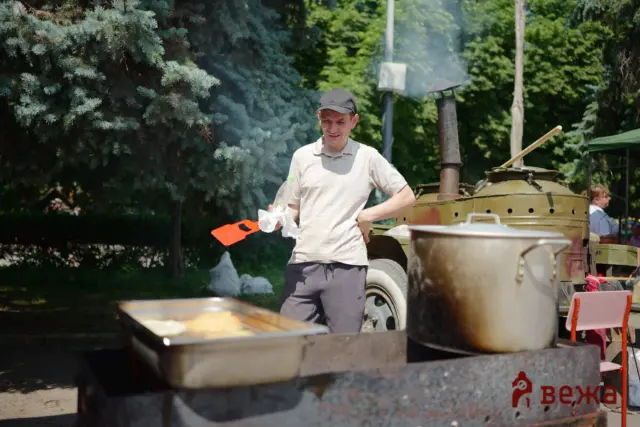 Man in a beige polo shirt and black cap stands next to a large outdoor smoker, smoke rising as he holds an orange spatula and a white cloth.