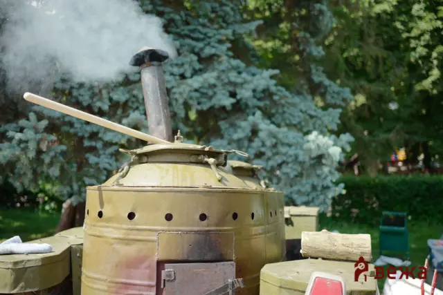 Close-up of a weathered steam boiler with a smokestack emitting steam, outdoors among trees with a wooden rod resting on top.