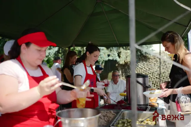 People in red aprons serving food at an outdoor market stall under a green canopy.