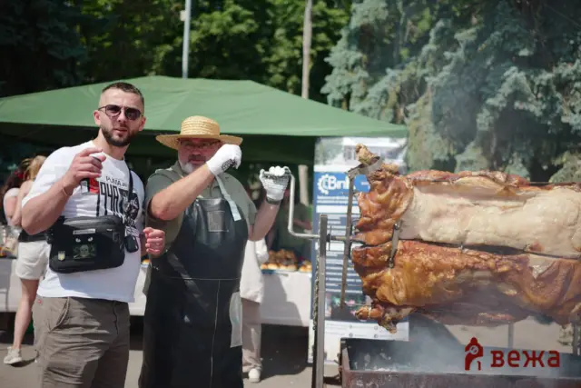 Two men at an outdoor barbecue, one wearing a straw hat, cooking a large roast on a spit