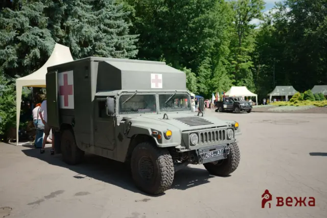 Military medical vehicle with red cross symbols parked at an outdoor event, tents and people in the background.