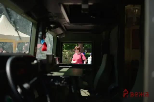 View through a vehicle window showing a woman in a pink shirt standing outside, holding papers, with another person nearby.