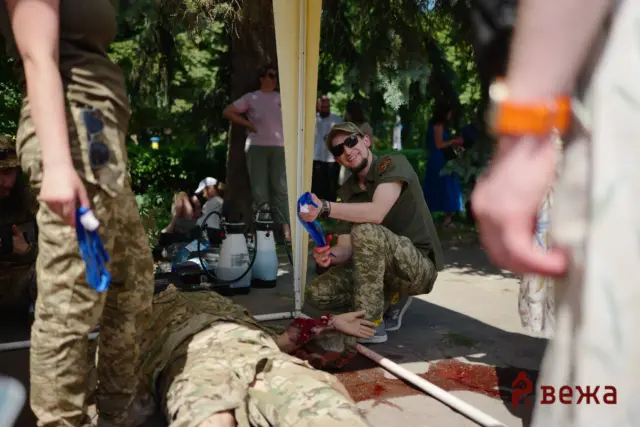 Outdoor military training scene with camouflage-clad soldiers; a mock casualty lies on the ground with fake blood near a hose and medical gear under a canopy.