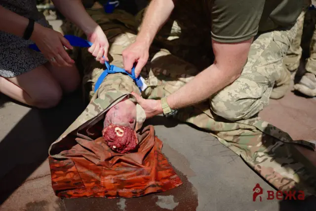 Military medics bandage a severely injured leg with an open wound and visible blood on a red cloth.