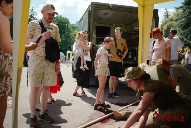Group of people outdoors under a yellow canopy near an open truck; a man kneels on the ground with a red liquid nearby as others watch.