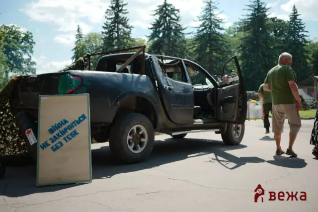 Damaged dark blue pickup truck with open doors at an outdoor scene; a man in a green shirt stands nearby amid trees and signs.