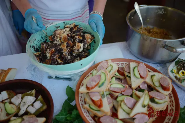 Hands in blue gloves assembling crostini with cucumber and sausage on a white tablecloth plate