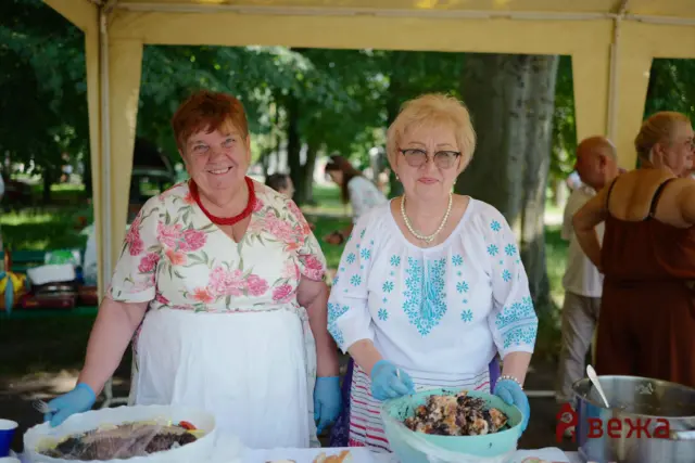 Two smiling women in floral blouses wearing gloves stand at a table with desserts under a yellow canopy at an outdoor event, serving food.