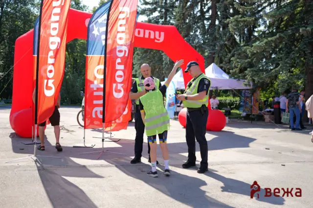 Public safety event outdoors with police officers talking to a child at a bright red inflatable arch and banners in the background.
