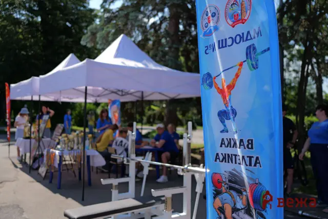 Outdoor event with white canopy tents, people at tables under trees, and a tall blue banner featuring a weightlifter illustration and Cyrillic text in the foreground.