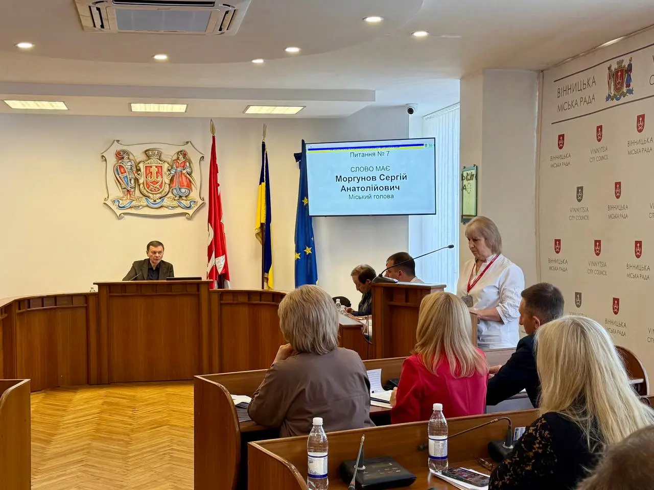 Municipal council chamber with a man at a curved wooden podium, flags behind him, and attendees seated at desks facing the speaker. A screen displays a name and title in Ukrainian/Soviet style setting.